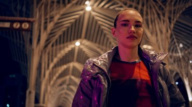 Mutlu havalı genç kız Gotik şehir gecesinde dolanıp duruyor. Yakın plan. Kameraya poz veren kendine güvenen şık kadın gece geç saatlerde modaya uygun ceket giyer. Kaygısız çekici esmer, özgür hissederek eğleniyor.