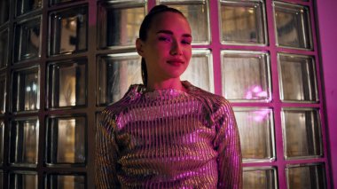 Cam duvarın önünde duran çekici bir manken. Kendine güvenen güzel bir kadın gülümseyen kamera. Parlak gümüş elbiseler giyen mutlu parti kızı portresi. Gece hayatı gece kulübü konsepti