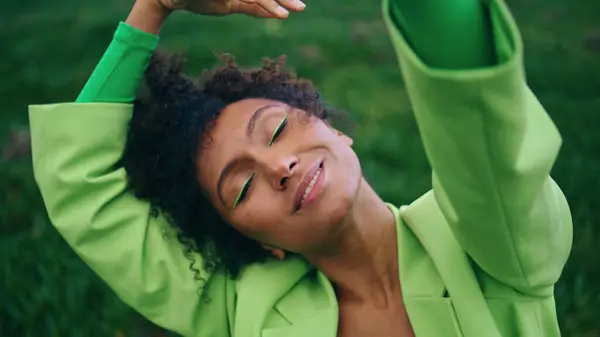 Inspired girl dancer dancing sensually on nature close up. Relaxed african american woman closing eyes enjoying dance gloomy evening at green grass. Curly lady in trendy bright suit dancing outdoors.