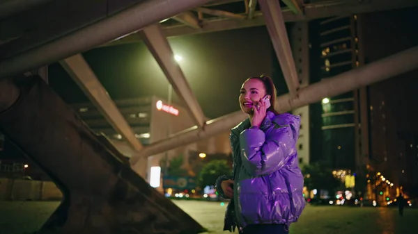 Carefree trendy woman calling phone standing urban street at night lights. Happy relaxed girl talking telephone outdoors. Cute cheerful brunette in jacket enjoy communication by modern smartphone.