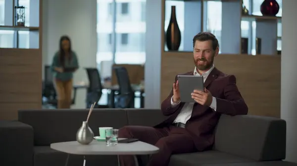 Smiling professional lawyer touching tablet computer screen at corporate cabinet. Cheerful bearded businessman holding modern pad sitting comfortable sofa. Happy manager financial office work concept.