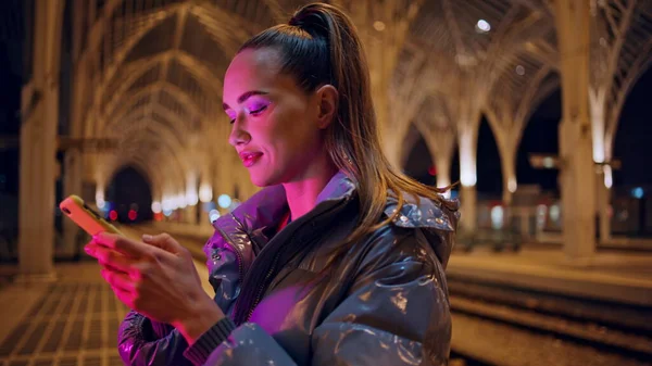 Cute girl looking smartphone screen standing at night city under beautiful gothic architecture close up. Pretty young woman reading message on phone smiling outdoors. Trendy tourist teenager concept.
