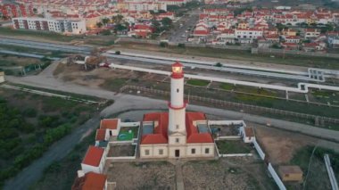 Deniz kenarı kasabasında kırmızı deniz feneri drone görüntüsü. Liman şehrindeki şehir altyapısı. Akşamları kiremitli çatı binaları olan kıyı yerleşim alanı. Petrol yakıtını depoya taşıyan Petrokimya boru hattı.