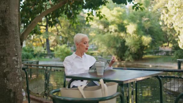 カフェテラスで新聞を持っている成熟した女性 夏の公園で休むリラックスした年金読書雑誌 金持ちのエレガントなビジネスマンがテーブルに座っているページを回します 退職した人のライフスタイル — ストック動画