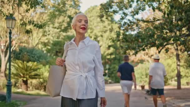 高齢の女性が日光の中で公園の道を歩いています 笑顔は 夏に都会の庭を散策する一人でリラックスした上級休憩 遊歩道を楽しむバッグを保持するスタイリッシュな灰色の髪の大人の 退職生活スタイルコンセプト — ストック動画