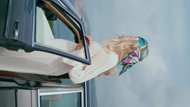 站在云彩海岸的女人开着门的车 迷人的金发年轻司机坐在前座复古汽车的垂直视频 在班杰明那的无忧无虑的女游客在海滨享受旅行 — 图库视频影像