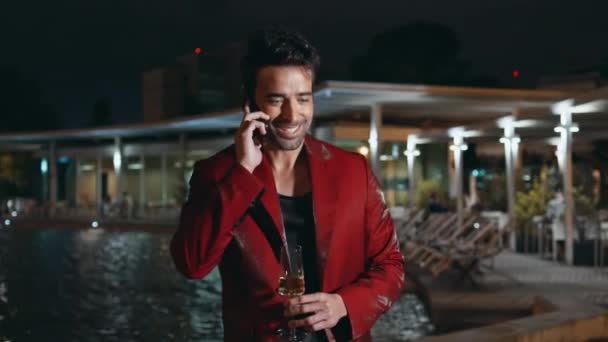 奢侈的商人结束了在湖边的晚会上庆祝的电话 迷人的西班牙人深夜在户外用智能手机进行特写 穿着红衣拿着香槟酒的帅哥 — 图库视频影像