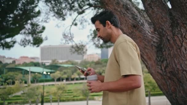 松懈的游客步行公园 手拿着饮料 拉近了距离 侧视迷人的他惊慌失措的家伙漫步在绿地上看智能手机屏幕 无忧无虑的男人享受周末喝咖啡的休息 — 图库视频影像
