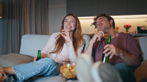 可爱的夫妇坐在舒适的公寓里看电影 在夜间特写 快乐的家人喜欢看电视 看啤酒瓶的叮当声 笑着快乐的夫妻一起在舒适的沙发上吃着薯片 — 图库视频影像