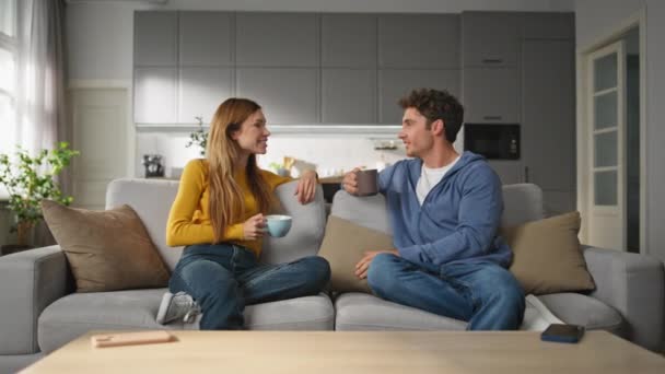 快乐的恋人在家里喝咖啡聊天 快乐的男朋友女朋友拿着杯子在客厅里笑 无忧无虑的家庭在舒适的公寓里玩乐 人的生活方式 — 图库视频影像