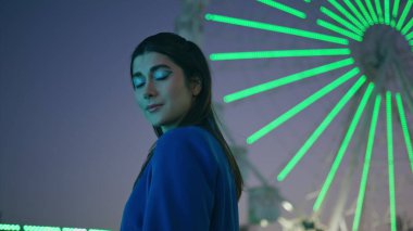 Aydınlatılmış lunaparkta dönme dolap pozu veren güzel bir model. Gülümseyen çekici kız kameraya bakıyor, gösterişli ve moda makyajlı. Hafta sonu açık hava tatili. Şehir gece hayatı konsepti