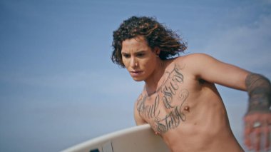 Kaykayda dengede duran mutlu kıvırcık kaykaycı, güneşli bir günde beyaz sörf tahtası tutuyor. Kaygısız gülümseyen kaykaycı yavaş çekimde hızlanıyor. Dövmeli yarı çıplak hipster aktif hobisinin keyfini çıkar.