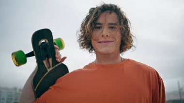 Havalı genç kaykaycı. Kapıya yakın sörf tahtasıyla yürüyor. Modern kaykayla bulutlu bir sokakta gezen çekici, rahat adam hippisinin portresi. Kıvırcık mutlu genç aktif bir hobiden hoşlanıyor