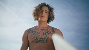 Ciddi modaya uygun sörfçü portresi güneşli gökyüzünün önünde beyaz bir sörf tahtası tutuyor. Çekici kıvırcık sörfçü yaz doğasına bakan kameraya bakıyor. Sportif dövmeli adam aktif hobisinin keyfini çıkar..