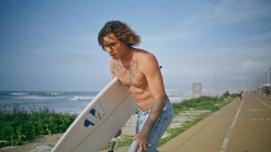 Deniz kenarında sörf tahtası tutan havalı dövmeli adam. Genç trend hippi paten seti üzerinde ilerliyor. Kıvırcık yarı çıplak kaykaycı hafta sonu sörf yapmak için sörf tahtasına biniyor..