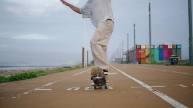 Adam bulutlu gökyüzünde kaykay pistine ayak basıyor. Kasvetli okyanus sahil yolunda kaykay antrenmanı yapan yakışıklı adam. Şık genç kaykaycı ağır çekimde uzun panoda en büyük hobisini yapıyor.