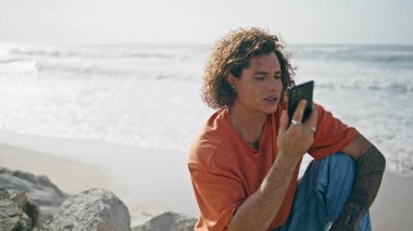 Okyanus sahilinde güneşli bir günde telefonu kullanan genç ve yakışıklı bir turist. Kıvırcık adam telefon görüşmesini bitiriyor. Yaz mevsiminde oturuyor. Yakın çekim. Gevşek dövmeli model akıllı telefonlu hafta sonunun tadını çıkar..