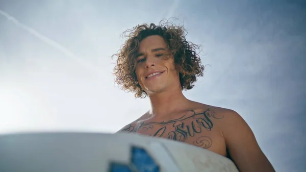 Attractive happy surfboarder standing in front beautiful blue sunny sky close up. Portrait of handsome extreme hipster man posing outdoors holding white surfboard. Curly tattooed man looking distance