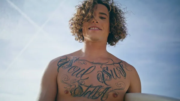 Handsome tattooed man surfer posing at sunny sky backdrop closeup. Portrait of attractive curly guy hipster holding white surfboard looking camera with smile. Carefree happy model enjoy active hobby.