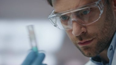 Biyoteknoloji araştırmacısı yenilikçi ilaçlarla test tüpüne bakıyor. Odaklanmış mikrobiyolog laboratuardaki tıbbi maddeleri kontrol ediyor. Güvenlik gözlüklü genç bilim adamı elinde uyuşturucu örneği tutuyor.