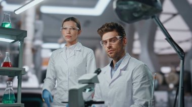 Yenilikçi klinik ekipmanlarında koruyucu gözlüklü iki tıp araştırmacısının portresi. Gelişmiş laboratuarda poz veren kendinden emin profesyonel bilim adamları. Kameraya bakan genç doktorlar.