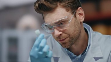Test tüpünü analiz eden üzgün laboratuvar araştırmacısı tatminsiz deney sonuçlarını yakından inceledi. Güvenlik gözlüklü bilim adamını hayal kırıklığına uğratmak araştırmada başarısız oldu. Eldivenli erkek doktor elinde ilaç örneği tutuyor..