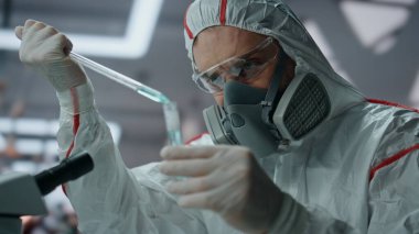 Koruyucu giysi bilim uzmanı test tüpünü kimyasal maddeyle dolduruyor. Tehlikeli madde maskeli ciddi bir bilim adamı biyokimya laboratuvarında deney yapıyor. Virüsle çalışan araştırmacı.