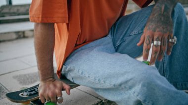 Yaz sokağında uzun sörf yapan genç kaykaycı kapanıyor. Çekici, kıvırcık erkek kaykaycı portresi modern kaykayla dışarıda dinleniyor. Mutlu stil sahibi hippi gülüşlü kamera