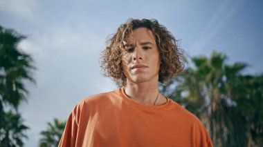 Ciddi yüzlü, kendine güvenen kıvırcık adamın portresi. Çekici genç hippi güneşli bir günde yeşil ağaçların önünde poz veriyor. Moda odaklı erkek modeli rahatla doğaya yakın dur.