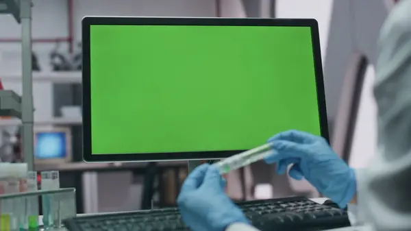 Chemist working at chroma key monitor in advanced laboratory close up. Unrecognizable scientist in gloves examining test tube at lab. Technician exploring vaccine looking green screen computer data