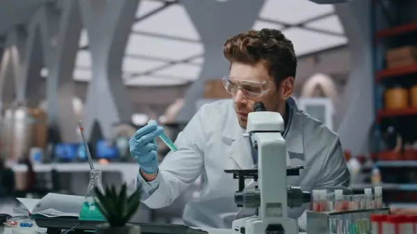 Disappointed researcher looking microscope in medical laboratory close up. Dissatisfied man scientist holding test tube feeling failure. Sad specialist in safety eyeglasses failed vaccine research.