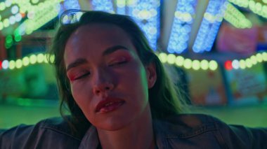 Gece lambalarında poz veren yakın model. Çekici kız neon ışıklı lunaparkta gözlerini kapayarak eğleniyor. Hafta sonu şehir karnavalında harika bir bayan kamera görüntüsü..