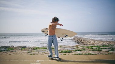 Güneşli bir günde elinde sörf tahtası olan dövmeli bir kaykaycı. Summerboardwalk plajında buz pateni numarası yapan harika bir sörfçü. Yarı çıplak adam okyanus sörfüne giderken ağır çekimde kayıyor.