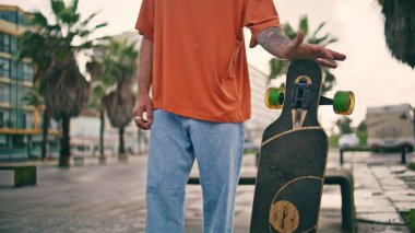 Kaykaycı eli kaykayla oynuyor. Açık havada yakın plan. Genç kaykaycı elinde dönen modern uzun kaykay bulutlu bir cadde tutuyor. Bilinmeyen trend gençlik ağır çekimde aşırı hobi yapmaktan zevk alıyor.