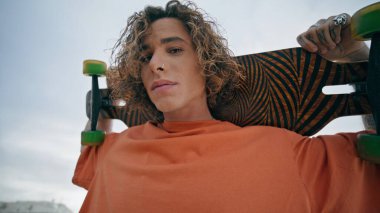 Bulutlu yaz manzarasında kaykay portresi tutan kıvırcık adam. Dışarıdaki kameraya bakan uzun kaykaylı çekici bir kaykaycı. Modayı takip eden hippiler sokakta tek başlarına eğleniyorlar.