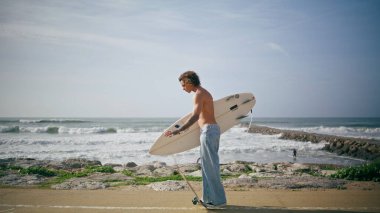 Genç kaykaycı elinde sörf tahtasıyla güzel okyanus dalgalarının yanında poz veriyor. Yarı çıplak, hippi bir adam modern kaykayla yol kenarında dikiliyor. Dövmeli adam deniz tatilinde hobisinin keyfini çıkar..