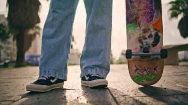 Ayakları genç kaykaycı kaldırımda dikiliyor ve kaykayı yakından tutuyor. Bilinmeyen profesyonel kaykaycı ağır çekimde asfaltta uzun tahta çeviriyor. Tanınmayan adam yaz güneşinde sokakta poz veriyor..