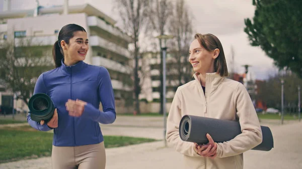 Sport ladies talking on walk at spring neighbourhood. Close up girls friends holding fitness mats going to yoga class together. Sporty happy women having fun laughing at workout break. Active hobby