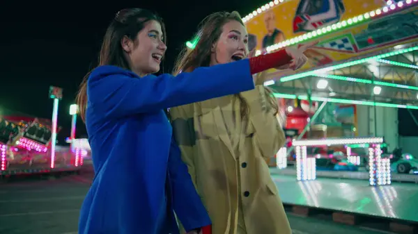 Cheerful friends having fun at night amusement park. Happy girls laughing walk illuminated carousel street together. Gorgeous excited girlfriends sisters talking enjoy summer weekend at city funfair