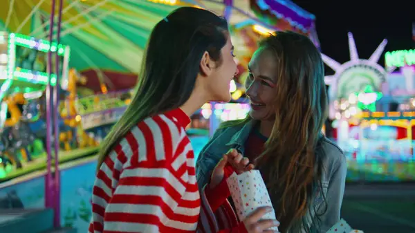 Joyful girls talking sharing secrets at luna amusement park. Happy best friends hold snacks laughing at illuminated funfair carousel. Cheerful excited teenagers having fun. Female friendship concept