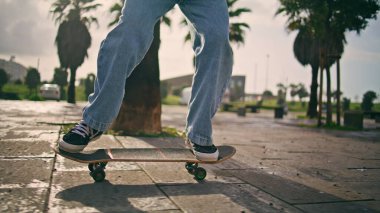 Güneşli şehir caddesinde kaykaya binen adam ayakları yakın mesafede. Bilinmeyen bir hippi, açık havada uzun panoda dengede duruyor. Erkek bacakları ağır çekimde asfaltta paten antrenmanı yapıyor. Genç sporcu ekstrem sporlardan hoşlanıyor.