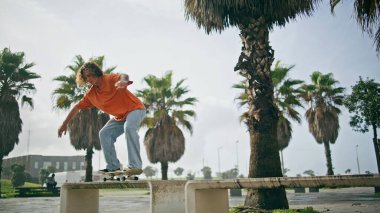 Genç aktif patenci güneşli tropikal caddede aşırı numaralar yapıyor. Profesyonel kaykaycı asfaltta banktan uzun kaykayla atlıyor. Maceracı hippi adam ağır çekimde eğitim hobisi