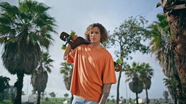 Güneşli şehir doğasında, omuzlarında kaykayla poz veren genç havalı patenci yaklaş. Yakışıklı, kıvırcık, hippi görünümlü, uzun pano tutan bir kamera. Moda adamı aktif yaz gününün tadını çıkarıyor..