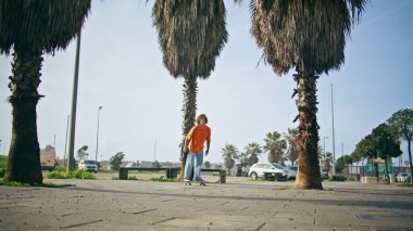 Genç maceraperest kaykaycı güneşli şehir caddesinde paten antrenmanı yapıyor. Palmiyelerin yanında uzun sörf yapan son moda profesyonel patenci. Aktif erkek hippi kaykay antrenmanında ağır çekimde dengede duruyor.