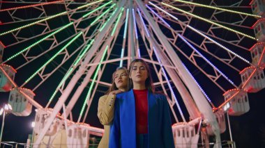 Kız arkadaşlar gece dönme dolap pozu veriyor. Geceleri yanıp sönen ışıkları aydınlatan iki model lunapark atlıkarıncasını dinlendiriyorlar. Güzel kızlar renkli festivalde kameraya bakıyor. Hafta sonu Luna Parkı.