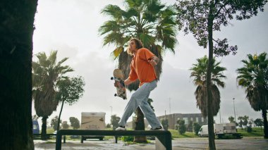 Güneşli caddedeki banktan kaykaya atlayan aşırı enerjik kaykaycı. Genç kaykaycı, tropik palmiyelerin yanındaki uzun tahtalarda profesyonel numaralar yapıyor. Ağır çekimde hobi yapmaya odaklanmış bir çocuk.