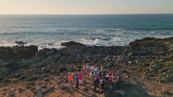 海岸に立っているグループの人々は 嵐のような美しい海洋無人機の景色を洗いました 美しい岩の海岸の夏の夕方に集まる会社の若者 沿岸の石が泡を作る景色の波がクラッシュ — ストック動画