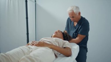 Klinik osteopat masaj yapan kadın kafa yakınlaştırması yapıyor. Odaklanmış adam hastane koğuşunda hasta yaralanmasını, iyileşme spazmlarını inceliyor. Rahatlamış Afro-Amerikan ziyaretçi fizyoterapist.