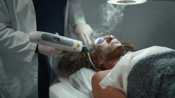 レーザーパルス患者の顔のクローズアップ サロンで皮膚を剥離するコシエティシャンの手 アフリカ系アメリカ人女性が再生手順に眼鏡をかけた 未知の皮膚科医は治療を持ち上げる — ストック動画