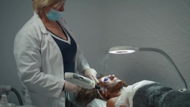 皮肤科医生在温泉里用碳面罩剥皮 戴眼镜躺在床上的非洲裔美国妇女人数激增 专业的化妆品师使用激光设备治疗面部皮肤 现代美容院 — 图库视频影像